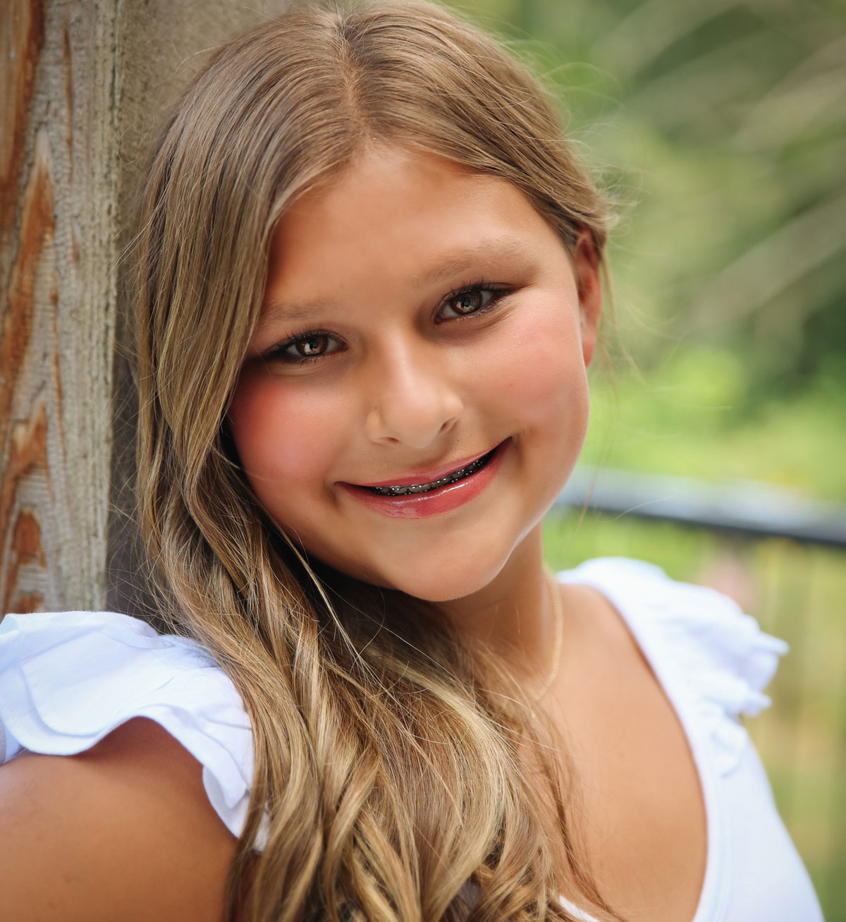 In this professional portrait, a white girl with long wavy hair leans against a wooden post outdoors. The background shows an out of focus natural setting.