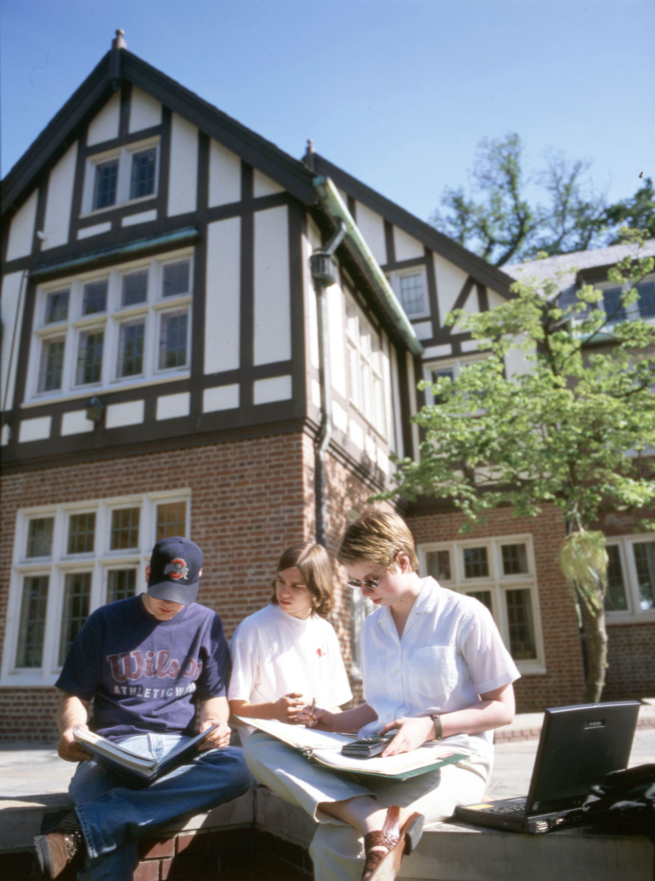 In a photo from 1999, three people study together in front of Kuhn House, a Tudor-style home converted into a building for academic use. The people are seated on a low brick wall, each holding notebooks or a laptop. The sun shines on them.