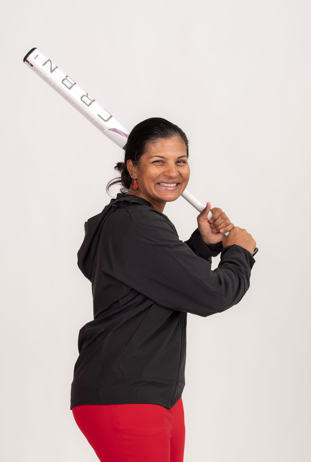 Kirin Kumar giggles as she poses in a batting stance, holding a baseball bat over one shoulder as if she’s getting ready for a pitch. 