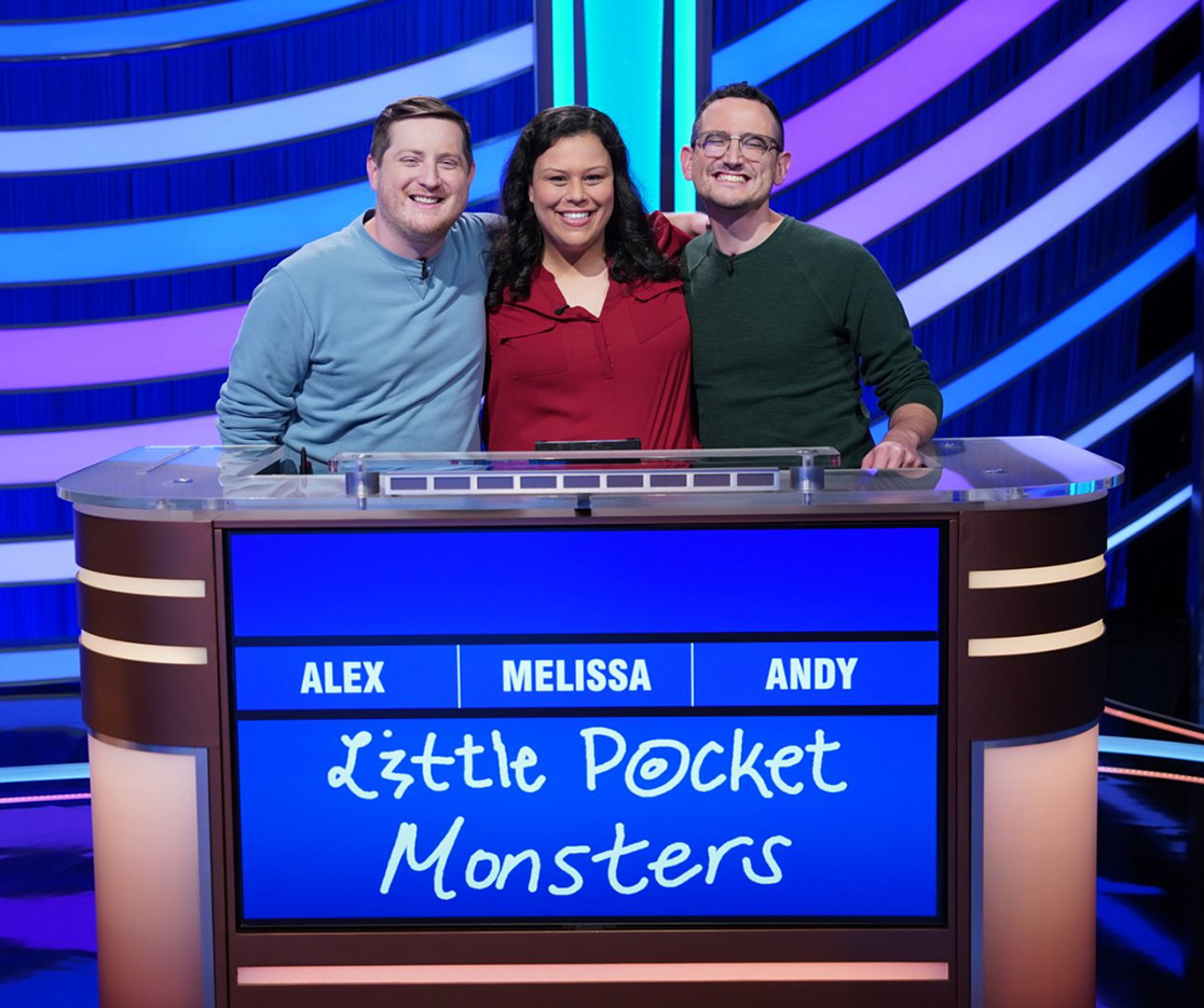 Three Buckeyes pose with their arms around one another on the stage of Jeopardy! The two on the sides are men; in the center is the woman. They seem genuinely close and pleased to be competing together.