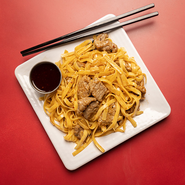 Flat, saucy noodles are shown mounded on a square plate with chunks of beef mixed throughout. A cup of sauce and a pair of chopsticks sit at the edges.