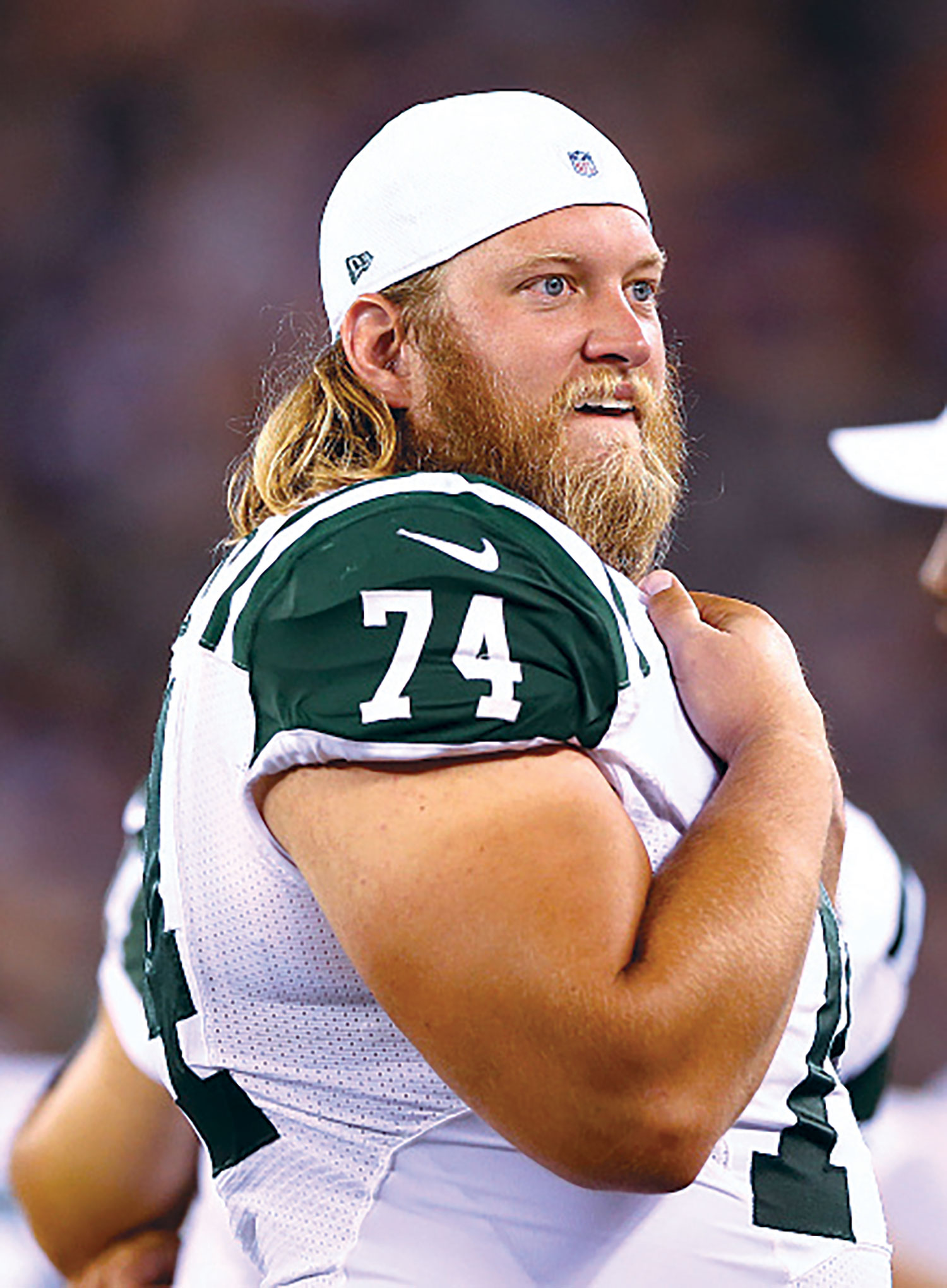 A football player with long blond hair stands along the field in a white and green uniform with the number 74, holding one arm across his chest pads. He is Nick Mangold and he looks pleased by what he’s watching.
