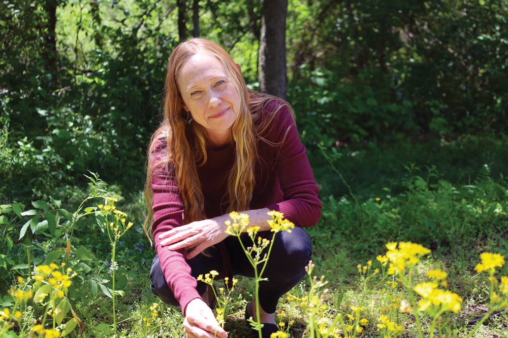 A professor with long hair looks into the camera with a knowing but peaceful expression as she crouches amid the prairie.