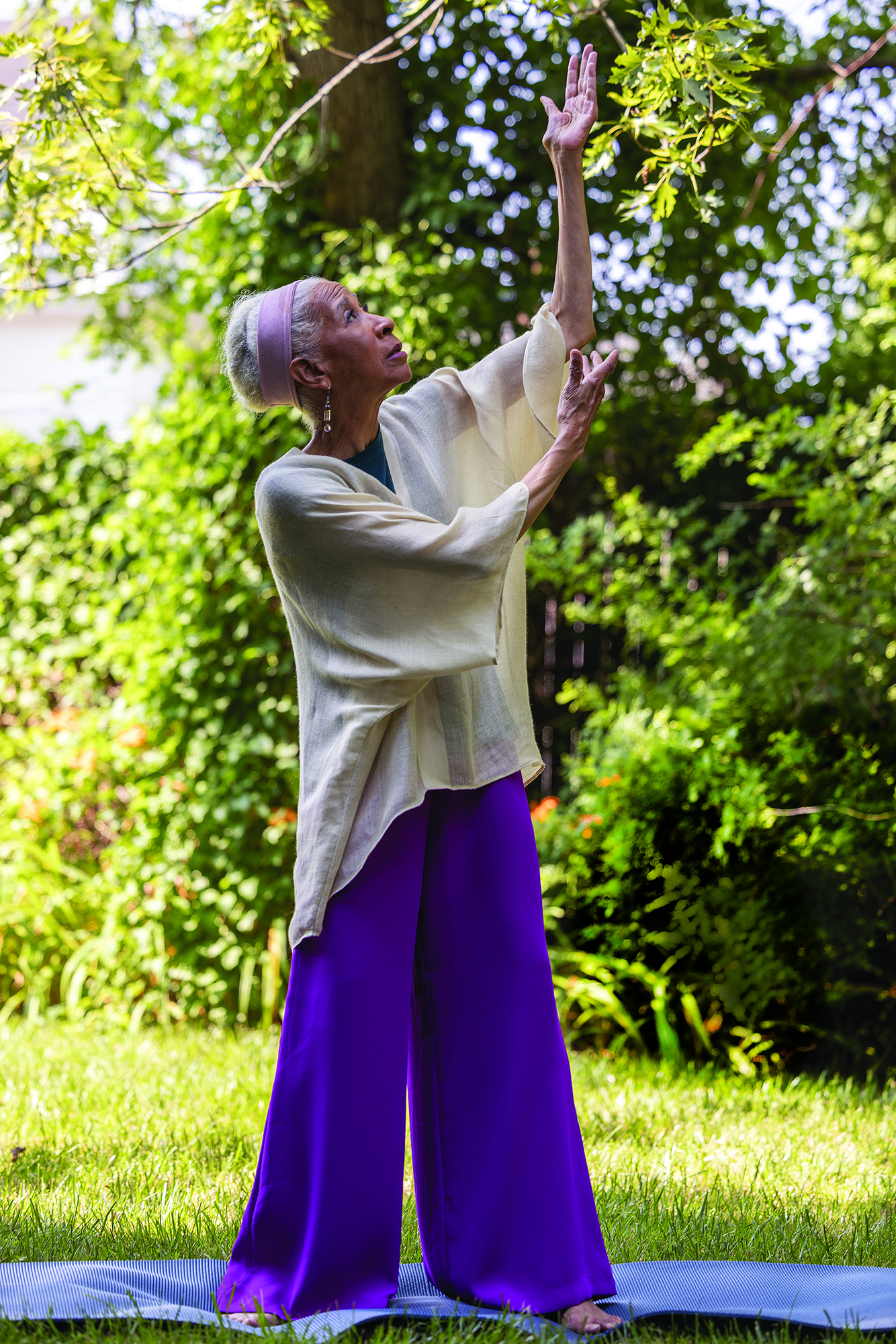 Dianne practices Qigong surrounded by trees and grass. Her arms are bent at the elbows a little more than 90 degrees and raised to her left. She looks up past her higher left hand. She wears flowey clothes and her expression is serene.