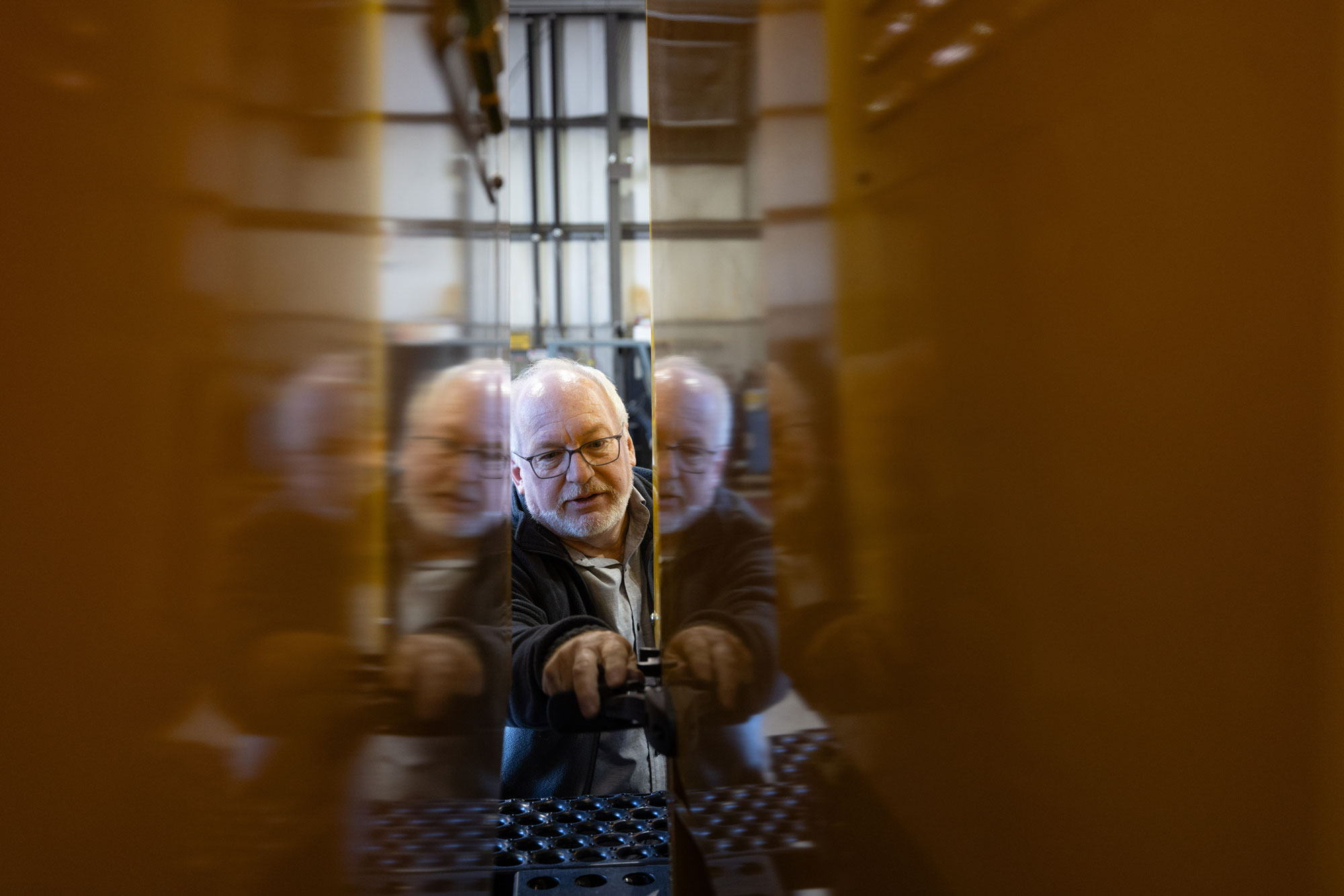 Dale adjusts equipment inside a narrow space between two large vertical panels. The scene is viewed through the gap between the panels, creating reflections of him on both sides. Machinery and structural elements are visible in the background.