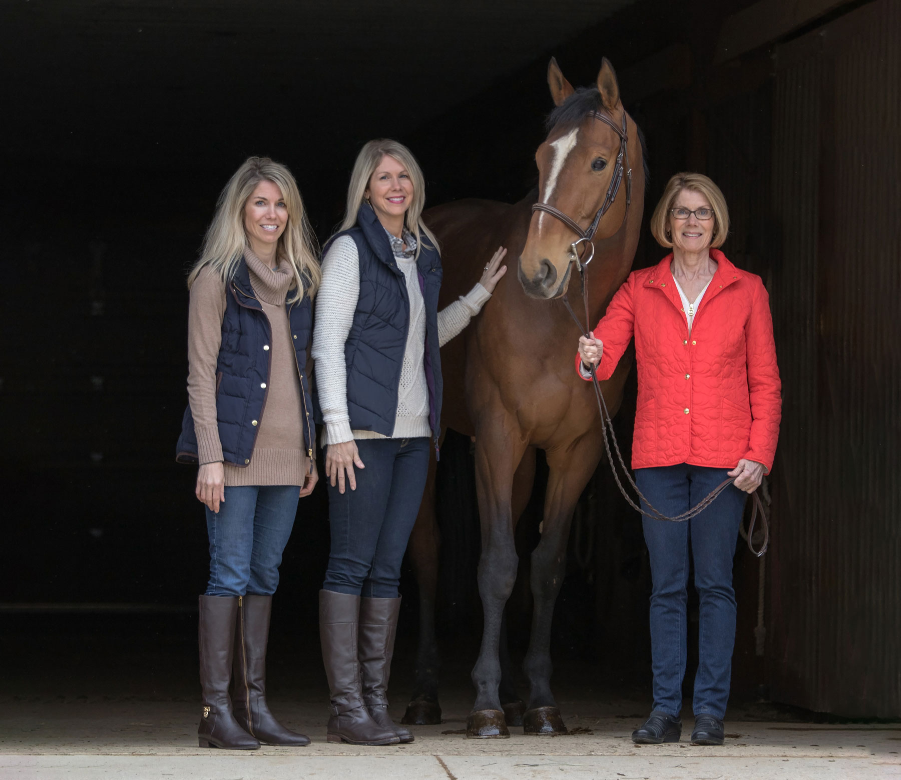 Three people stand beside a horse inside a barn entrance, with the mom holding the horse’s lead rope and her two grown daughters standing on the other side of the horse. They all smile as they pose.