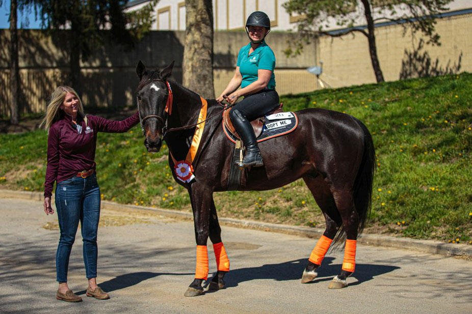 A person sits on a saddled horse wearing protective riding gear, while Winnie Morgan Nemeth stands beside the horse with one hand on the horse’s neck. The horse wears leg wraps and has award ribbons and a sash across its chest. 