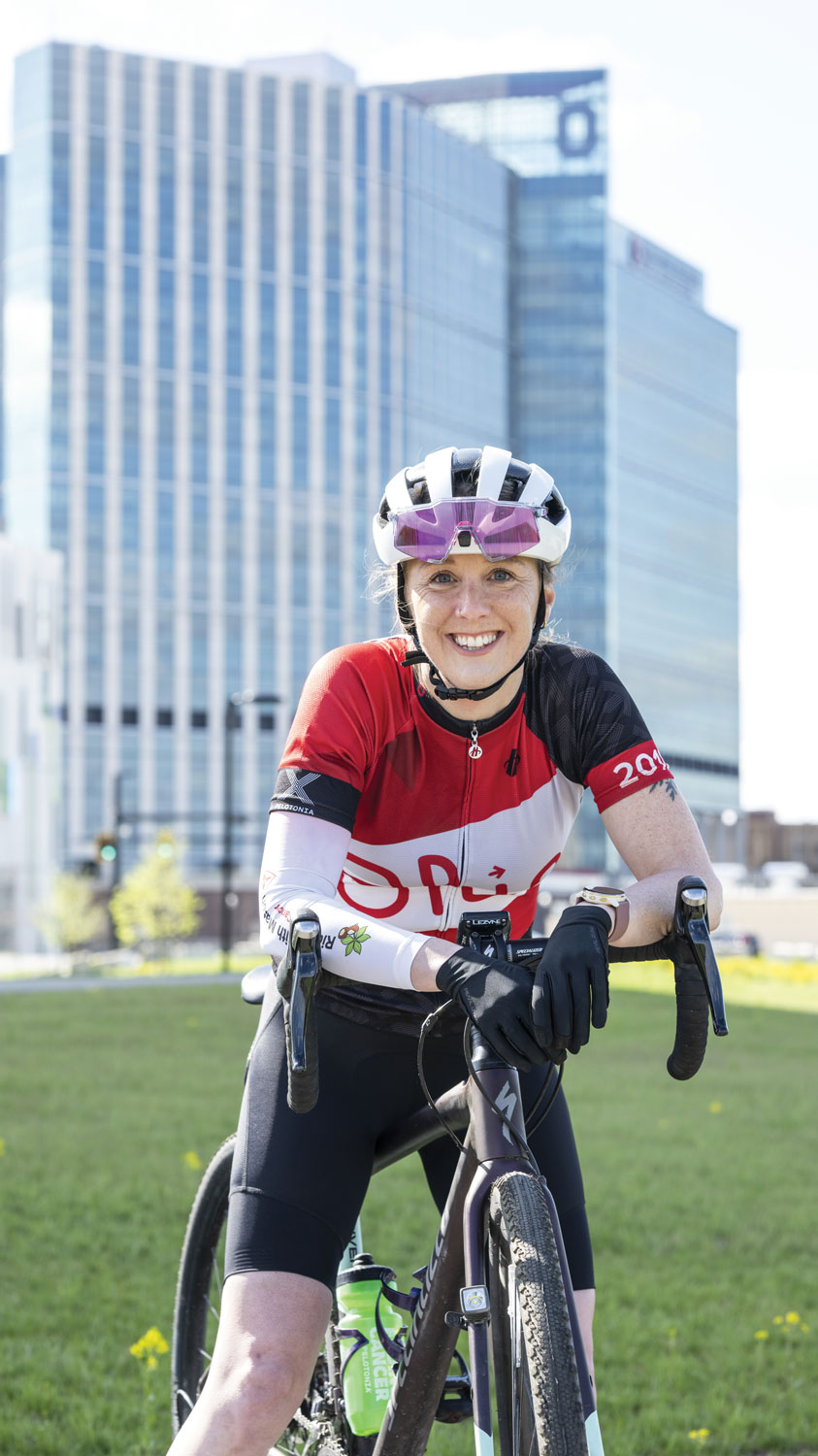 A woman on her bicycle and wearing a riding helmet and jersey smiles as she poses for a photo in front of Ohio State's new hospital tower