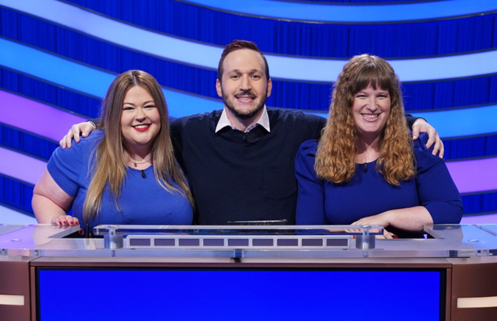 Three people, two women and a man, smile as they pose on the Jeopardy! set. The Buckeye is at right, and she has a big smile and long curly hair.