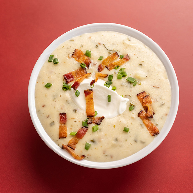 A thick, creamy soup is shown in a bowl, topped by a generous dallop of sour cream, sliced green onions and chopped strips of bacon.