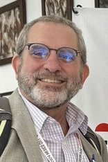 A man appears friendly and smart as he smiles while wearing glasses, a jacket and a backpack. 