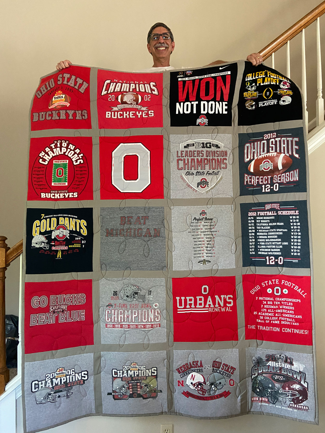 A mustached man stands on the tsairs of his home proudly displaying a quilt made from old Ohio State T-shirts