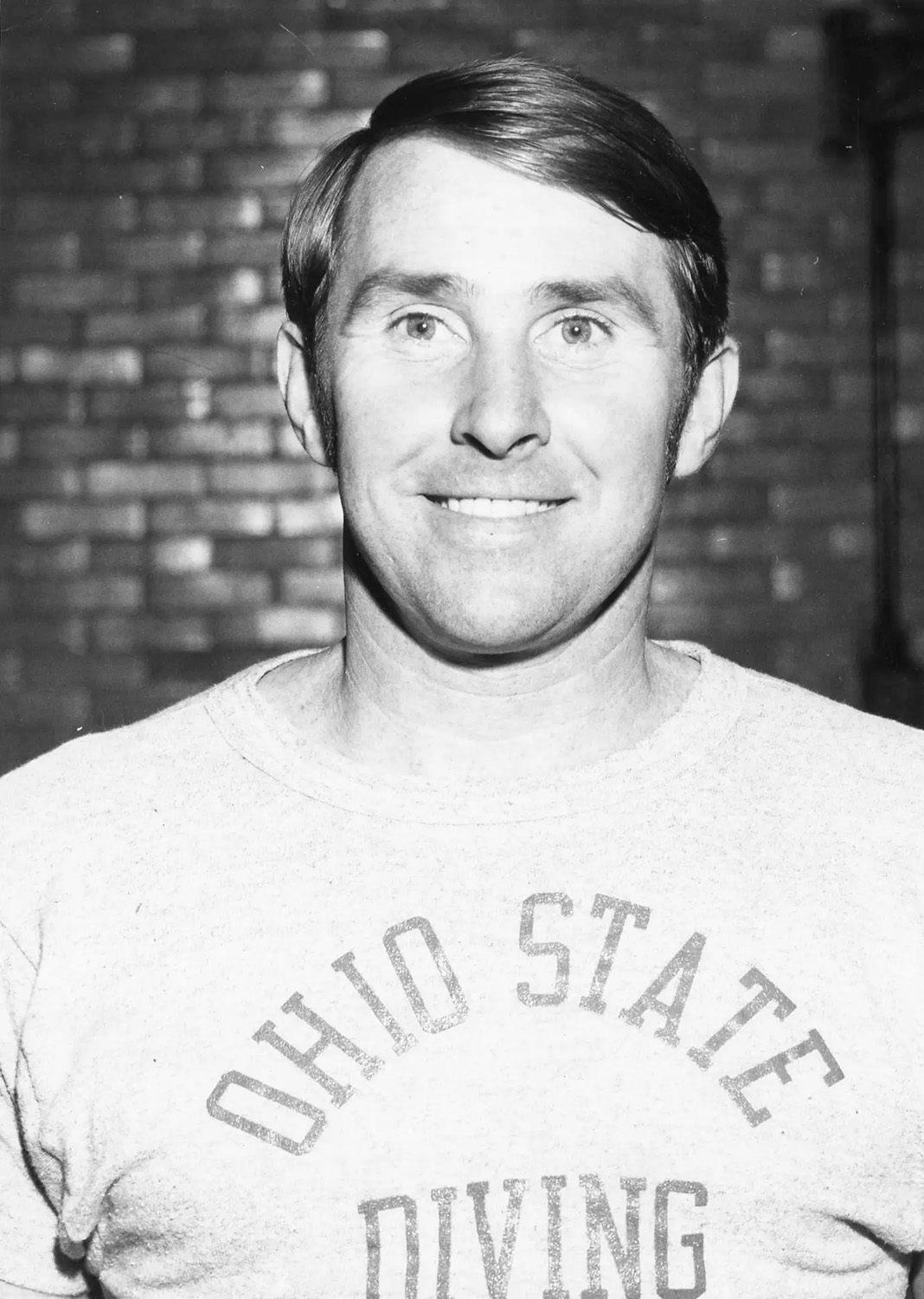 A clean-cut young man, Ron O'Brien smiles for the camera. This is a black and white photo, and he as eyes wide open and wears and ohio state T-shirt.