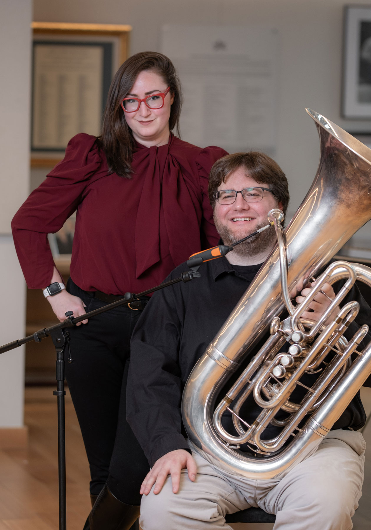 A young man smiles as he sits holding a tuba behind a microphone stand. His teacher stands close beside him with one hand on her hip. She wears bright red glasses and smiles with closed lips. She looks knowing and approving. They’re inside a music practice room.