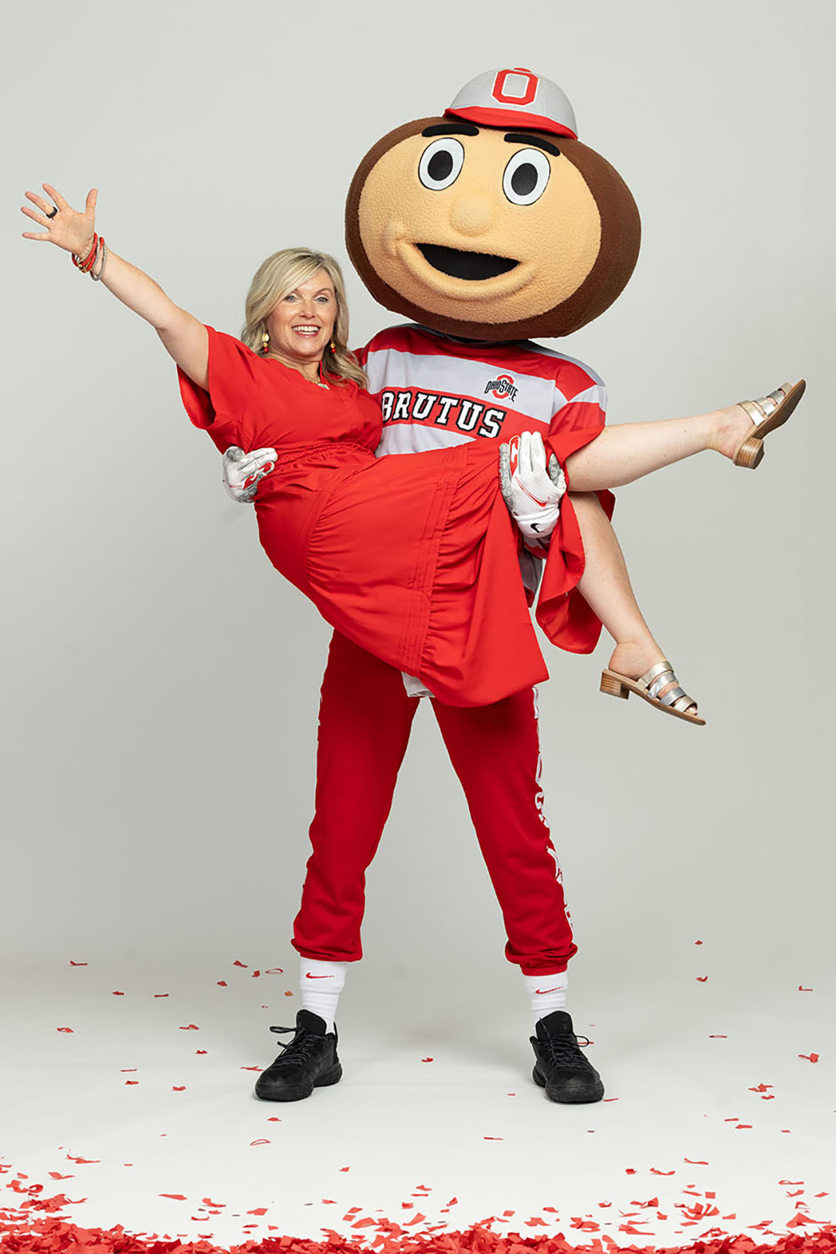 Tracy Stuck, a blond-haired white woman with a big smile and a scarlet dress, laughs as Brutus Buckeye hoists her in a cradle hold.