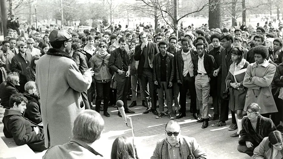 Black and white photo of crowed gathered around a Black man speaking with his back to the camera, wearing winter coat, glasses, and leather beret