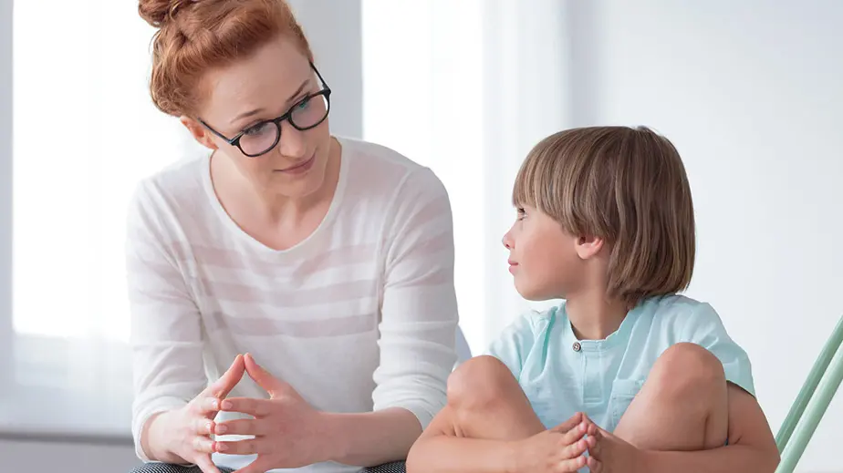 A woman with red hair and glasses is leaning on a table next to a young child.