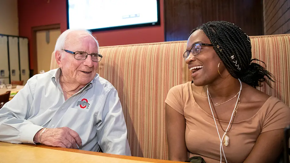 Cal is sitting with a young woman in a restaurant booth, laughing and smiling.