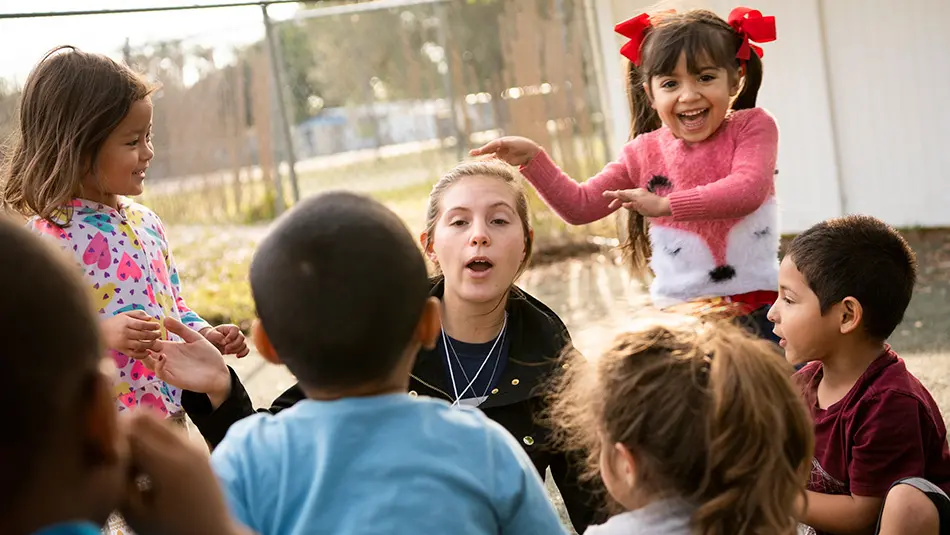 Lauren is seated playing duck duck goose with a group of children while a young girl with pigtails and red bows taps her on the head.