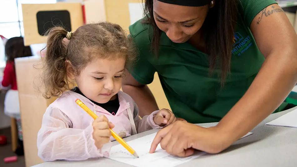 A young preschool girl is writing with a yellow pencil as an instructor leans in to point to her paper.