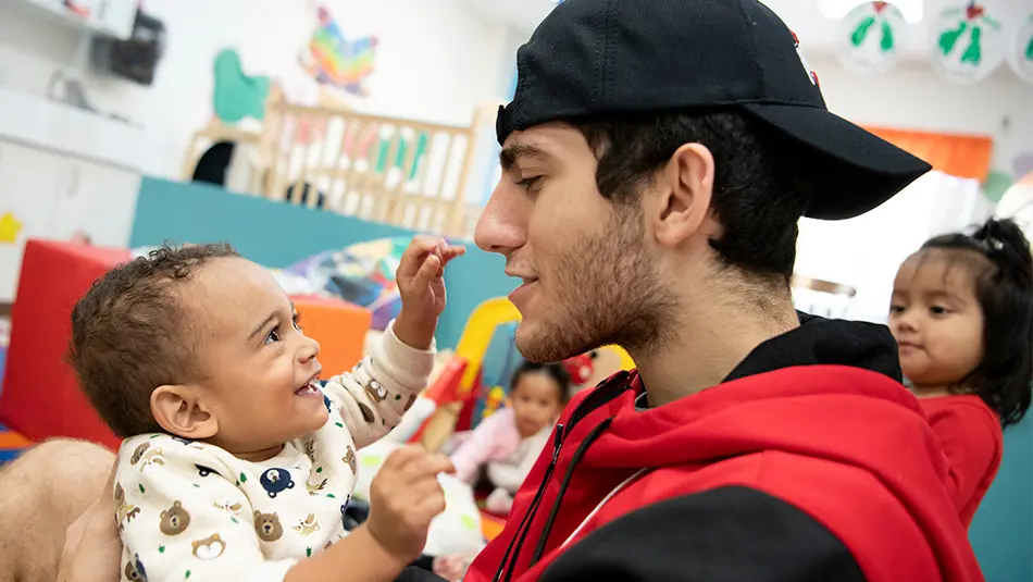 Kareem is holding a 1-year-old girl as she smiles and looks up at him during daycare.