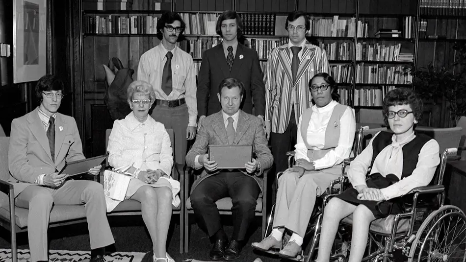 Black and white archival photo of a group of men and women in front of a bookshelf. There are 3 men standing behind a front row of seated men and women. 2 of the seated women are using wheelchairs.