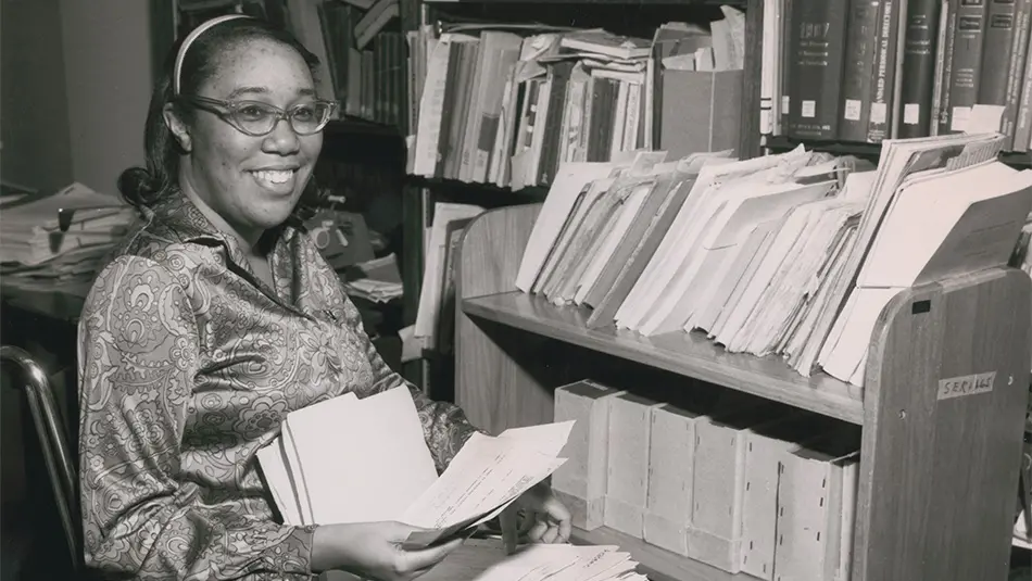Black and white archival photo of Jean Williams organizing papers in a library. She is using a wheelchair and is positioned next to a rolling shelf full of paper and binders.