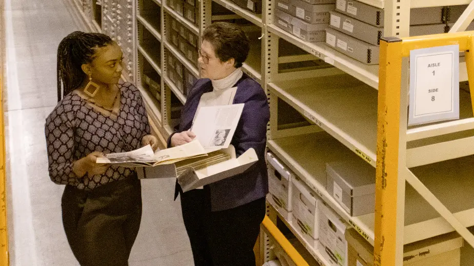 Two women are having a discussion in an aisle between tall metal shelves in a warehouse.