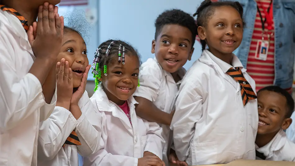 6 young students are standing near a low table top during a science lesson. All are wearing white lab coats.