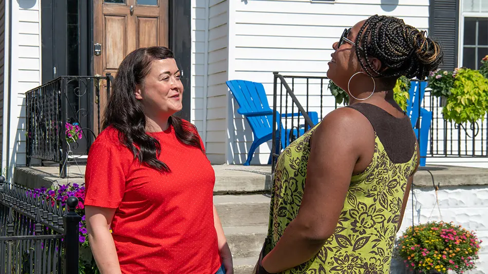 A woman wearing a red shirt and jeans is talking to her neighbor, another woman in a green printed dress.