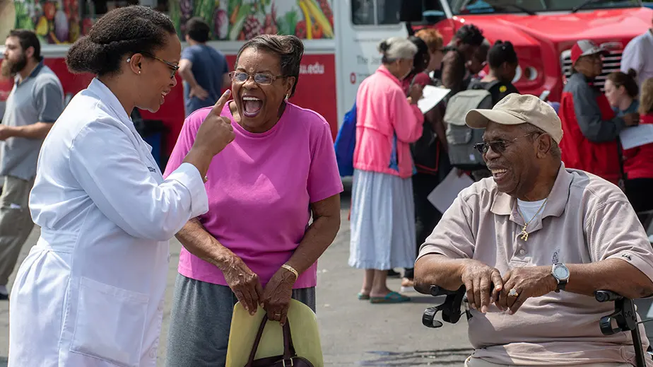 Dr. Sophia Tolliver is wearing a white lab coat while speaking to 2 of her patients. The patients are a woman in a pink shirt and a man in a tan ball cap and shirt who is seated.