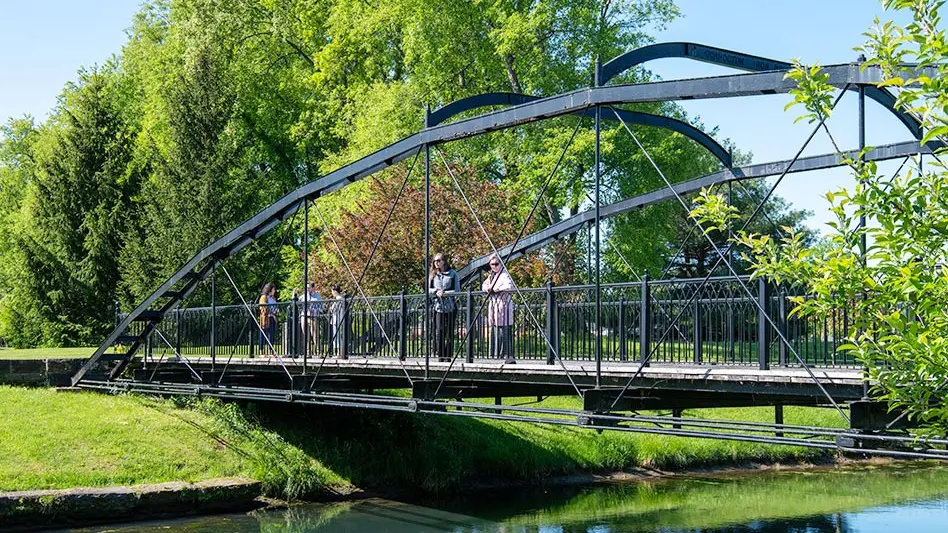 A black metal arched bridge spans a small river in a park