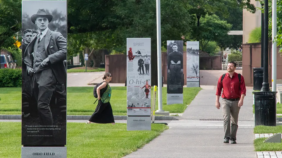 A man is walking along a sidewalk past tall freestanding installations of photos of historical OSU moments