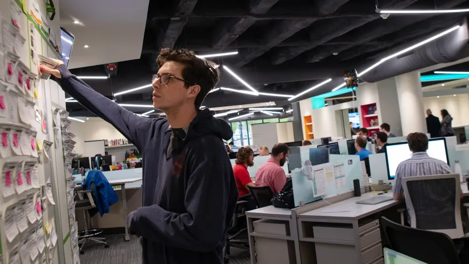 Paul is standing next to a large white board wall full of sticky notes. He is working in an office full of cubicles that can be seen behind him.