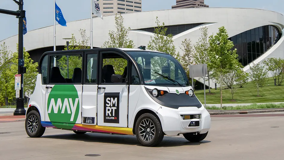 A self-driving shuttle is parked in front of a building in downtown Columbus. The shuttle looks like an extended Smart Car.