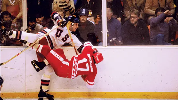 An action shot from the Miracle on Ice hockey game. A hockey player from team USA is standing on one leg after checking a player from the Soviet Union team who is falling to the ice.8