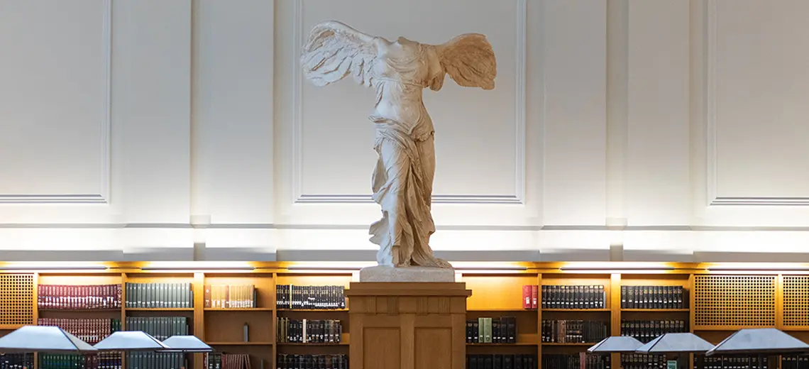 A statue titled "Nike of Samothrace" is on top of a pedestal in a library. The statue is a white marble representation of the goddess Nike with wings, no arms and no head remain.