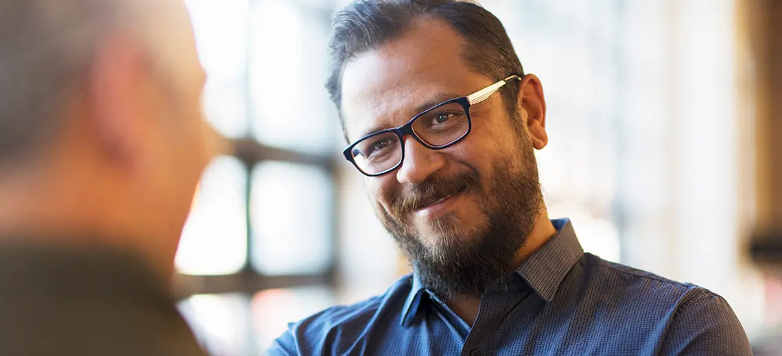 Man with beard and glasses smiling at blurry face. Sun coming in through window behind him. 