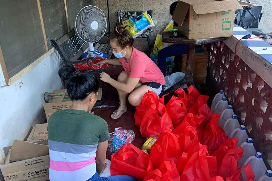 Two young women on a patio filling cardboard boxes, 3 rows of red bags, and a row of gallon jugs. There is a bench behind them with a fan.  