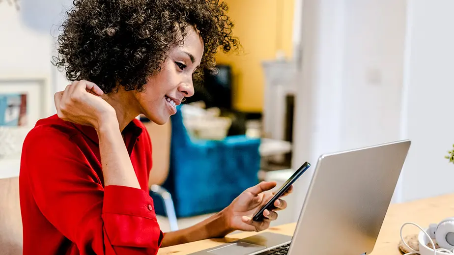 Woman with curly brown hair wearing a red long sleeve blouse is sitting in front of a laptop with a smartphone in her left hand.