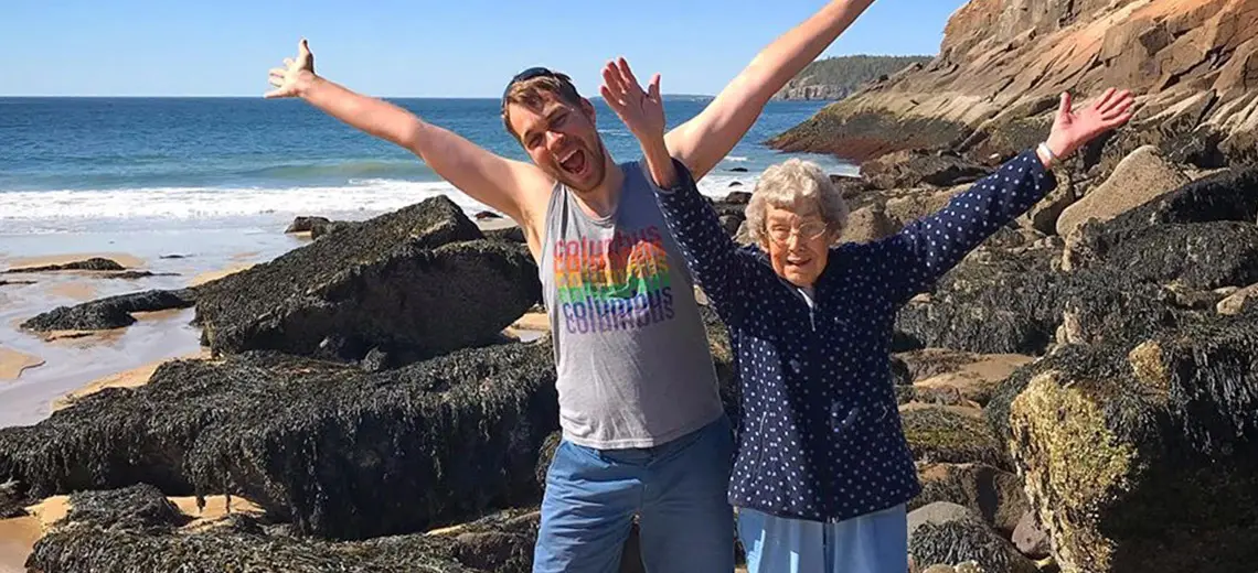 Brad and his Grandma Joy are standing next to one another on a rocky beach. They both are smiling and have both arms raised 