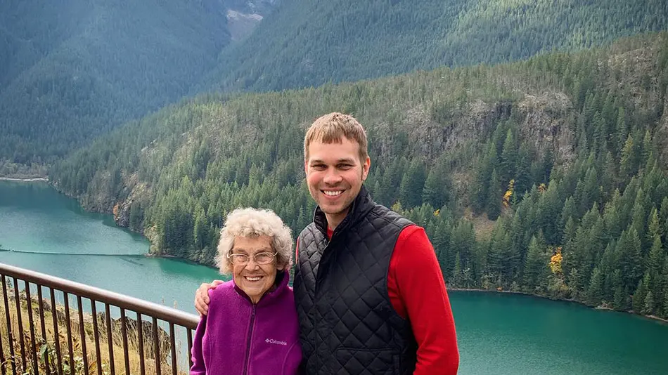 Brad is standing with his arm around Grandma Joy and they are both smiling. There is a lake and mountainside view behind them.