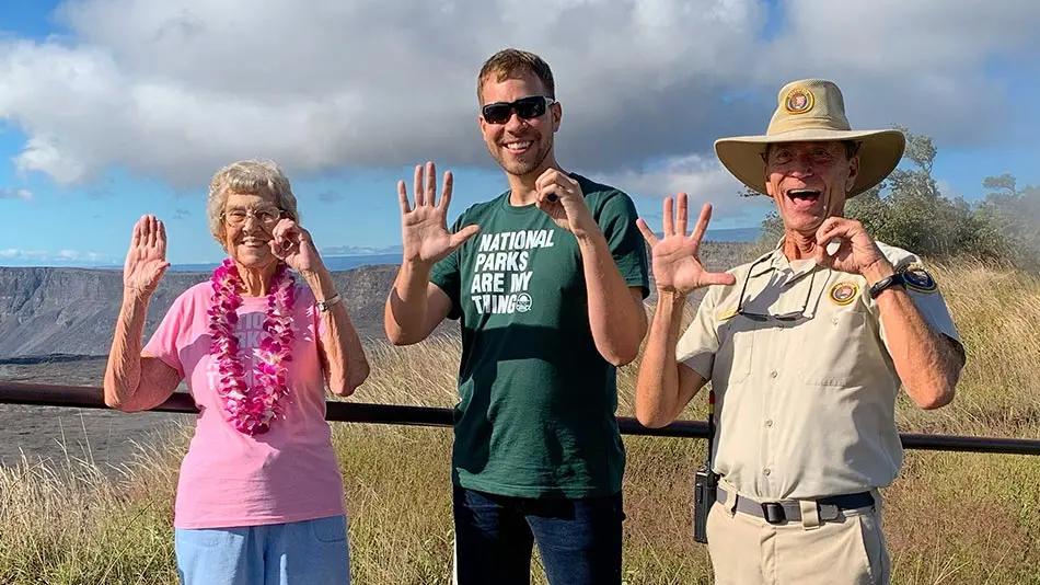 Brad and Grandma Joy are standing next to a park ranger. They all have their hands raised while making a 5 and a 0 with their fingers. 