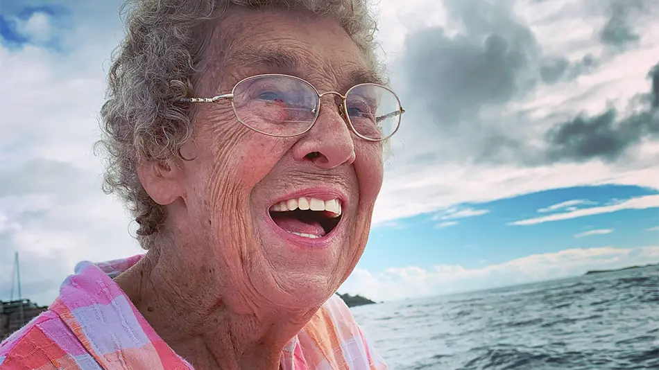 A close up of Grandma Joy smiling while on a boat at sea. She is wearing gold glasses and a pink and orange checkered shirt