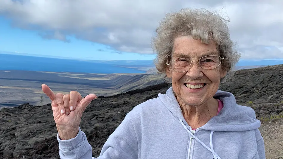 Grandma Joy standing in Hawai’i Volcanoes National Park wearing a gray hoodie. She has her hand raised with her thumb and pinky finger extended.