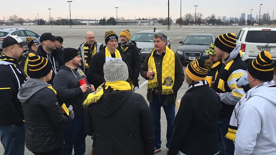 A large group of men and women are standing in a parking lot outside of Mapfre Stadium. Everyone is wearing black and yellow Columbus Crew attire.