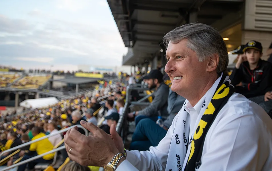 Edwards is clapping while watching the Columbus Crew soccer match inside Mapfre Stadium. He is smiling and wearing a white long sleeve shirt and a black and yellow Crew scarf.