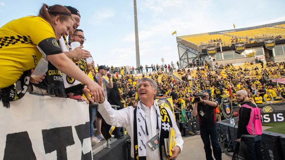 Edwards is walking along the stands of Mapfre Stadium and reaching up to shake hands with a fan in a yellow Columbus Crew shirt.