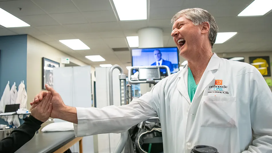 Edwards is standing in a physical therapy room wearing green scrubs and a white lab coat. He is reaching out with his right hand to grab someone's hand as he laughs.