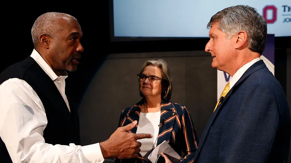 Gene Smith, Ann Fisher, and Pete Edwards are standing and speaking in front of a large projector's screen and a dark background